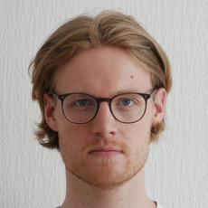 Blonder Mann mit mittellangem Haar vor weißer strukturierten Wand, Porträt von vorne.